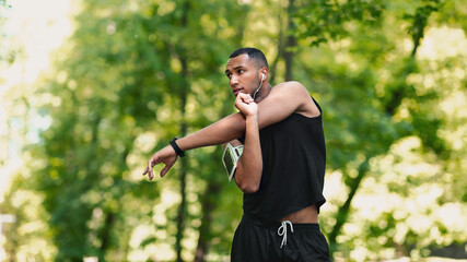 Cool black guy with earphones stretching his arm and listening to music on his morning workout at park, empty space