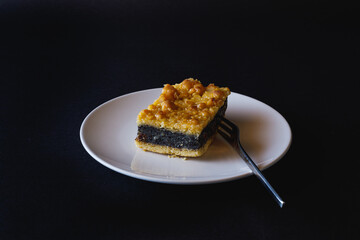 Slice of poppy seed cake on white plate with  dessert fork against a black background