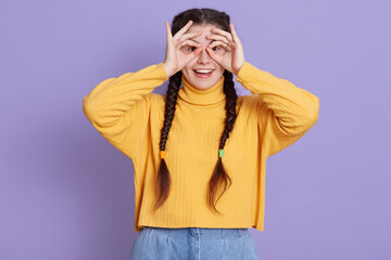 Fototapeta premium Excited brunette female with two pigtails standing with happy expression and covering her eyes with ok signs, making binocular with her fingers, looks at camera and laugh.