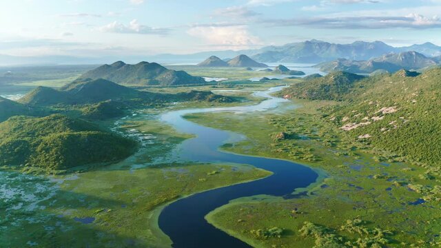 Blue river flowing through green valley toward distant mountains. Bends and curves of Crnojevica river in Montenegro, meandering through the marsh among hills on its way to lake Skadar. Aerial footage