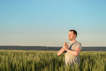 Naklejka na ściany i meble Adult man stands on a field in tall grass doing yoga during sunset. Digital detox, connection with nature concept. Copy space.