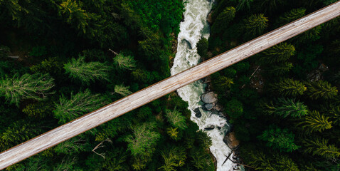 Top aerial view of rope walkway through the treetops in coniferous forest. Bird's eye view of hanging bridge crossing raging river, suspension bridge above green forest in the gorge of mountains © BullRun