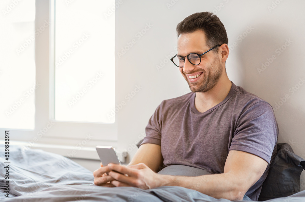 Poster smiling man using smartphone app sitting in bed at home