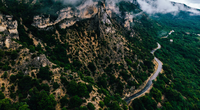 Aerial Scenery View Of Asphalt Road Pass In Rocky Mountains With Green Vegetation Covered With Fog After Rain. Bird's Eye View Of Rocky Hills With Picturesque Bike Path Above Which Hung Heavy Clouds