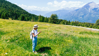 Aerial view of female tourist with backpack standing on green grass meadow and using smartphone for navigation route. Hipster girl wanderlust recreating in National Park during summer vacations