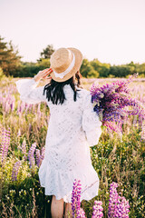 Girl in a hat with a bouquet of flowers. A girl in a white dress, in a straw hat, with a bouquet of lupins stands with her back facing the sun in a floral field, holding the hat