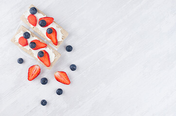 Organic season fruit sandwiches on dry brown grain crispbread with fresh ripe strawberry slices, blueberry, sweet cream cheese on white wooden table, top view, copy space.