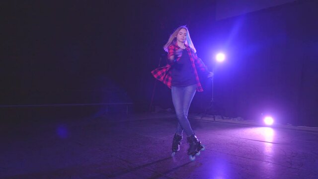 Beautiful young blonde girl spinning on rollers in one place with crossed legs on front wheels in dark scene of abandoned building under light of spotlights in slow motion. Concept of active lifestyle