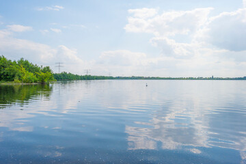 Edge of a sunlit lake with small islands with birds breeding reflecting a blue cloudy sky in summer