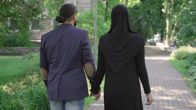 Back view of romantic muslim couple walking in park and talking. Camera follows loving bearded man in suit and young woman in hijab strolling along the alley on sunny summer day. Dating, love, romance