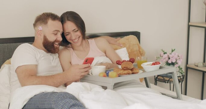 Young family and looking at smartphone using funny apps while spending time together in bed. Smiling woman and man relaxing and scrolling social media news feed while having breakfast .