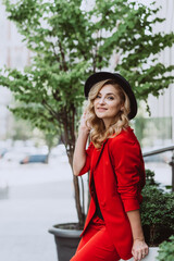 Obraz premium Young beautiful blond woman in a red suit and black hat stands outdoors in the yard next to the building. Stylish people. Soft selective focus.