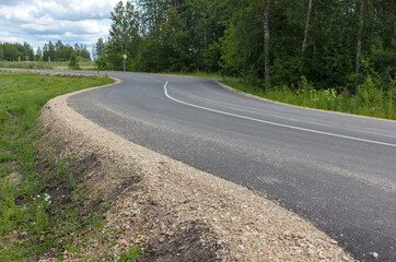View to a winding new asphalt road in a rural area. Gravel roadside, ditch for rainwater. Latvia. 