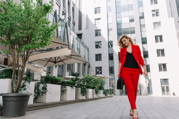 Young business woman blonde in a red suit is walking outdoors in the courtyard of a business center. Stylish people. Soft selective focus.