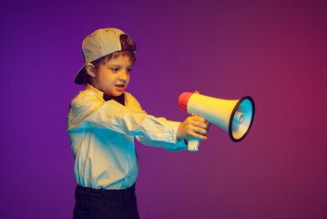 Shouting with megaphone, emotional. Joyful caucasian boy's portrait on dark studio background in neon light. Beautiful curly model. Concept of human emotions, facial expression, sales, ad, childhood.