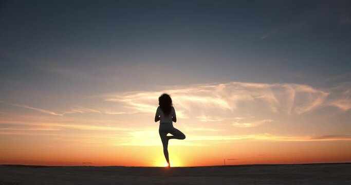 Against the backdrop of the bright sunset sky, the girl performs yoga asanas, raising her closed palms to the sky, first bends her right leg, and then grabs her left leg with her right foot and