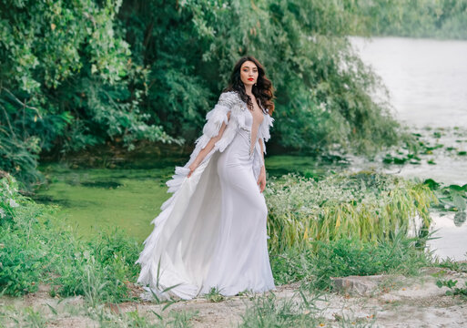 Beautiful Queen. charming woman bride walks on coast lake. Wedding outfit white long sexy dress, luxury cloak cape bird swan feathers. Brunette girl wavy loose hair. Backdrop summer nature green trees