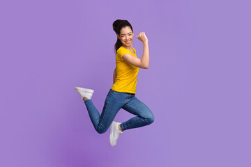 Overjoyed asian girl jumping with raised fists over purple studio background