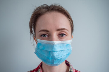 Portrait of woman wearing medical face mask, looking at camera in room with grey wall at home - close up front view. Self isolation, prevention, quarantine, COVID19, coronavirus, safety concept