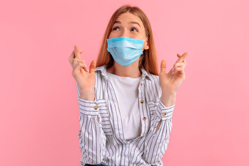 Focused girl in a medical protective mask on her face, fingers crossed for good luck, on a pink background