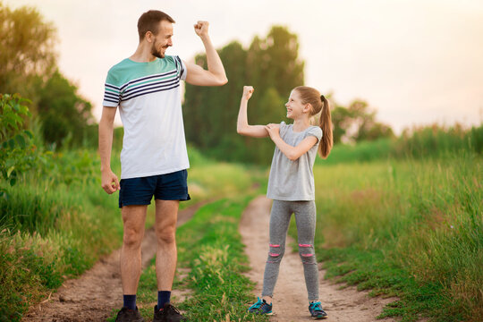 Full length portrait of handsome father and his cute little daughter showing their muscles, looking at camera and smiling at sunset