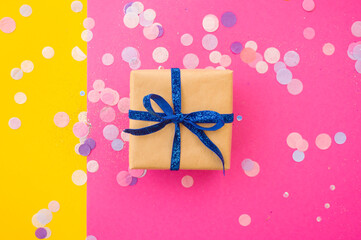 Giftbox tied with blue color ribbon on bright pink, pastel yellow background with colorful confetti and glitter. Festive background.