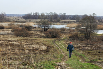 Naklejka premium A girl in an autumn jacket, hat and with a backpack takes a walk on nature in early spring. He looks at the vast expanses and enjoys the view.