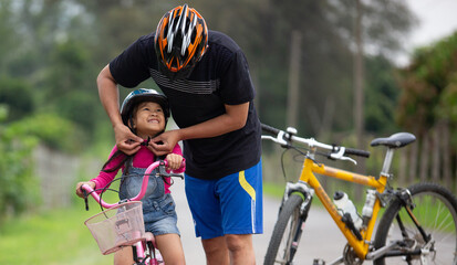Happy father and daughter cycling in the park wears a bicycle helmet to his daughter, togetherness...
