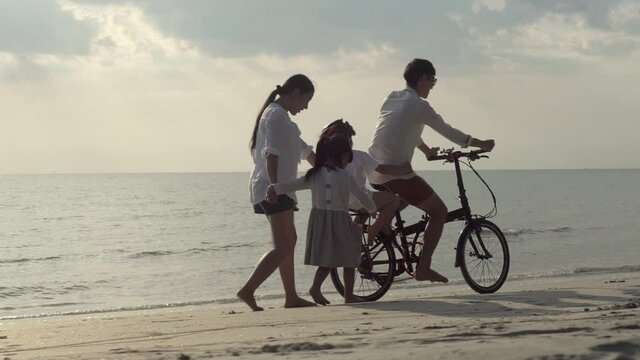 Happy asian family with father and son riding bicycle, mother and daughter walking on beach in vacation together, parent and children go to travel trip with cycling for leisure in summer.