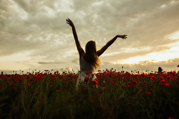 Silhouette of woman who stands toward the amazing sunset in a red poppy field with arms spread out. Summertime. Copy space.