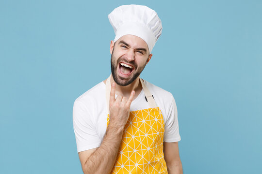 Crazy Young Bearded Male Chef Or Cook Baker Man In Apron White T-shirt Toque Chefs Hat Isolated On Blue Wall Background Studio. Cooking Food Concept. Depicting Heavy Metal Rock Sign, Horns Up Gesture.