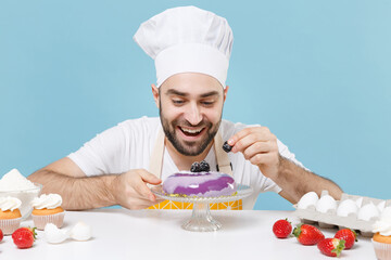 Funny male chef or cook baker man in apron white t-shirt toque chefs hat cooking at table isolated on blue background. Cooking food concept. Mock up copy space. Decorating dessert cake with berries.
