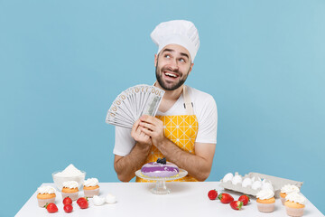 Pensive young bearded male chef or cook baker man in apron white t-shirt toque chefs hat cooking at table isolated on blue background. Cooking food concept. Hold fan of cash money in dollar banknotes.