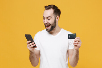 Excited young bearded man guy in white casual t-shirt posing isolated on yellow wall background studio portrait. People lifestyle concept. Mock up copy space. Hold credit bank card using mobile phone.