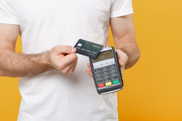 Cropped image of young man guy in white casual t-shirt isolated on yellow background. People lifestyle concept. Hold wireless modern bank payment terminal to process and acquire credit card payments.
