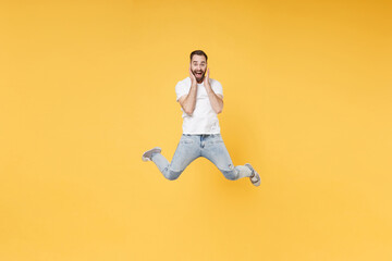 Fototapeta na wymiar Surprised young bearded man guy in white casual t-shirt posing isolated on yellow background studio portrait. People lifestyle concept. Mock up copy space. Jumping spreading legs put hands on cheeks.