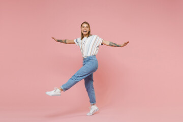 Fototapeta na wymiar Laughing young blonde woman girl in casual striped shirt posing isolated on pastel pink background studio portrait. People lifestyle concept. Mock up copy space. Dancing spreading hands rising leg.