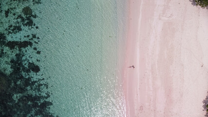 Aerial view of young woman sitting on pink beach. Turquoise sea color and pink sand beach. Copy space provided.
