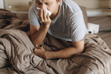 Young man with a cold blowing her runny nose with tissue. Asian man get sick sneezing from flu. Healthcare and medical concept. - obrazy, fototapety, plakaty