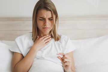 Sick young woman girl lying in bed with white sheet pillow blanket spending time in bedroom at home. Healthy lifestyle ill sick disease treatment concept. Hold thermometer, put hand on throat neck.