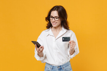 Smiling young brunette business woman in white shirt glasses isolated on yellow background . Achievement career wealth business concept. Mock up copy space. Using mobile phone hold credit bank card.