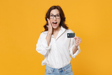 Excited young brunette business woman in white shirt glasses isolated on yellow background. Achievement career wealth business concept. Mock up copy space. Hold credit bank card, put hand on cheek.