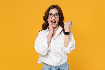 Excited brunette business woman in white shirt glasses isolated on yellow background. Achievement career wealth business concept. Mock up copy space. Wearing smart watch on hand, put hand on cheek.
