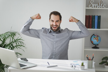 Cheerful young bearded business man in gray shirt sitting at desk work on laptop pc computer in light office on white wall background. Achievement business career concept. Showing biceps, muscles.