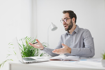 Excited young bearded business man in gray shirt glasses sit at desk in light office on white wall background. Achievement business career concept. Work on laptop pc computer hold pen spreading hands.