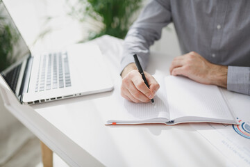 Cropped image of young business man in classic gray shirt sitting at white desk working on laptop pc computer in light office. Achievement business career concept. Writing notes in notebook.