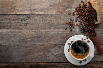 Top view above of Black hot fresh coffee with smoke and milk foam in a white ceramic cup with coffee beans roasted in burlap sack bag on wooden table background. Flat lay with copy space.