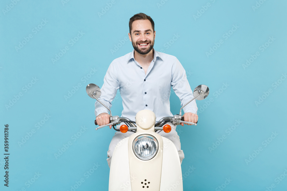 Wall mural Smiling young bearded man guy in casual light shirt driving moped isolated on pastel blue wall background studio portrait. Driving motorbike transportation concept. Mock up copy space. Looking camera.