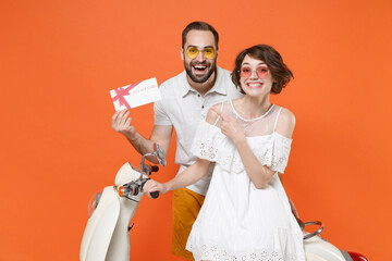 Excited young couple friends guy girl in white summer clothes glasses sit on moped isolated on orange background. Driving motorbike transportation concept. Pointing index finger on gift certificate.