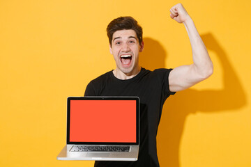 Screaming young man guy in casual black t-shirt isolated on yellow wall background. People lifestyle concept. Mock up copy space. Hold laptop pc computer with blank empty screen, doing winner gesture.